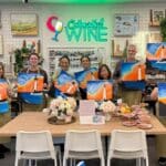 A group of smiling adults holding up their lighthouse paintings during a Colourful Wine Paint and Sip session, standing around a wooden table set with snacks and flowers in a cozy, art-filled studio café.