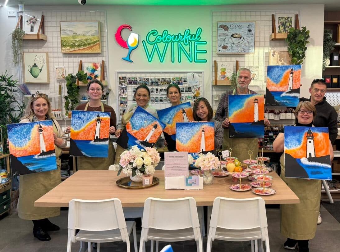 A group of smiling adults holding up their lighthouse paintings during a Colourful Wine Paint and Sip session, standing around a wooden table set with snacks and flowers in a cozy, art-filled studio café.
